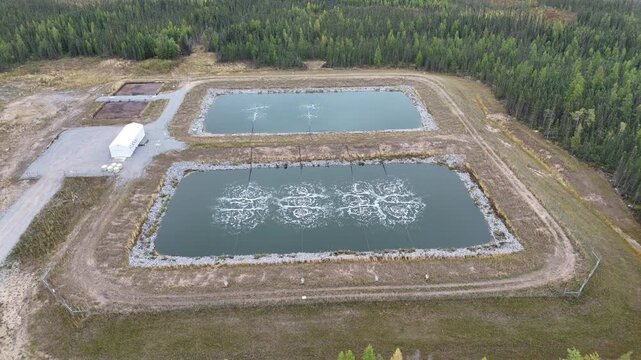 Aerial drone ascends and pulls away from wastewater lagoons near Ilford War Lake, northern Manitoba, Canada. Aeration bubbles, berms, gravel road, and boreal forest surround the site.