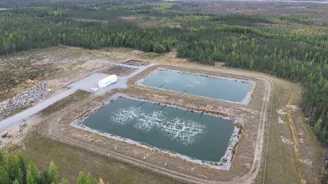 Aerial drone orbits left while moving toward wastewater lagoons near Ilford War Lake, northern Manitoba, Canada. Aeration bubbles, berms, gravel road, and boreal forest surround the site.