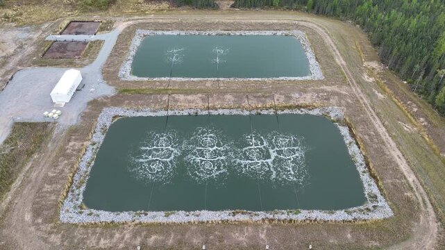 Aerial drone ascends while focusing on wastewater lagoons near Ilford War Lake, northern Manitoba, Canada. Aeration bubbles, berms, gravel road, and surrounding boreal forest visible.