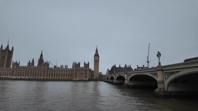 Iconic houses of parliament and big ben with westminster bridge over the river thames on a cloudy day in london, revealing the south bank embankment while moving backwards