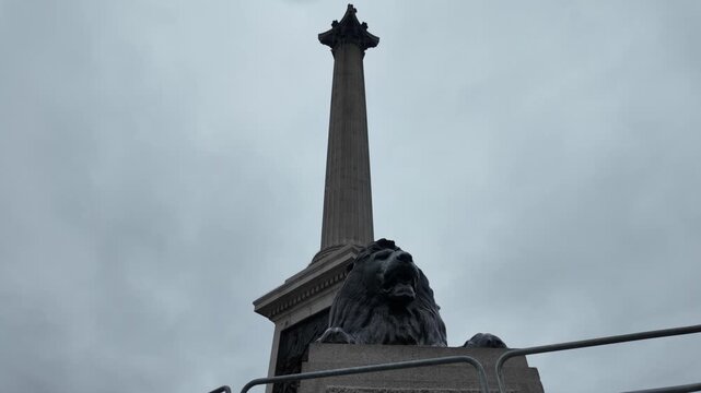 Nelson's column with a lion statue in trafalgar square on a cloudy day in london, revealing the famous monument from a moving point of view with a gray sky background