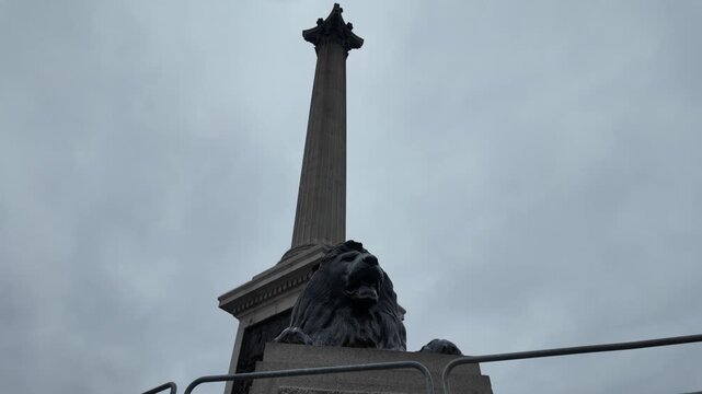 Nelson's column in trafalgar square featuring the iconic lion statue under a dramatic, cloudy sky, captured from a unique passing perspective on a classic double-decker bus