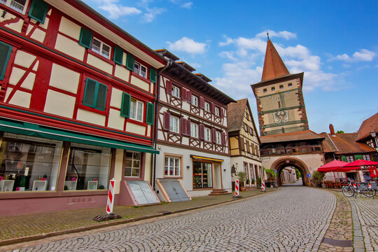 Colorful historic half timbered houses along cobbled street in Gengenbach old town with tower gate and flowers under blue sky