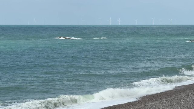 Waves rolling on the shore at Etretat Beach in France with wind turbines in the background during a clear day