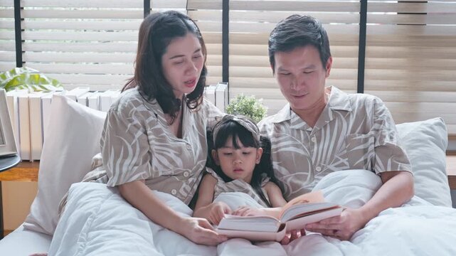Asian family bedtime routine with loving father and mother reading a storybook to their young daughter under a cozy blanket.