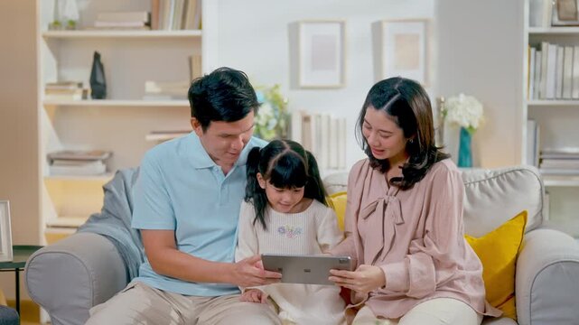 Asian family father, mother, and young daughter sitting on sofa and looking at digital tablet for education or entertainment