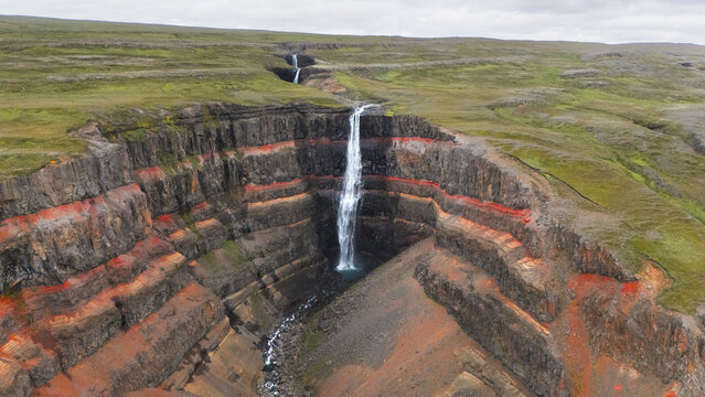 Aerial view of Hengifoss waterfall plunging down the cliff face, cutting through layers of vibrant red clay contrasting with the green landscape, Hengifoss, Iceland.
