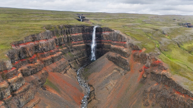 Aerial view of a cascading waterfall plunging dramatically through layered cliffs of dark basalt rock and vibrant red clay under a cloudy sky, Hengifoss, Iceland.
