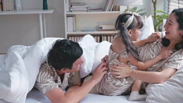 Cheerful Asian family tickling young daughter playing together on the bed