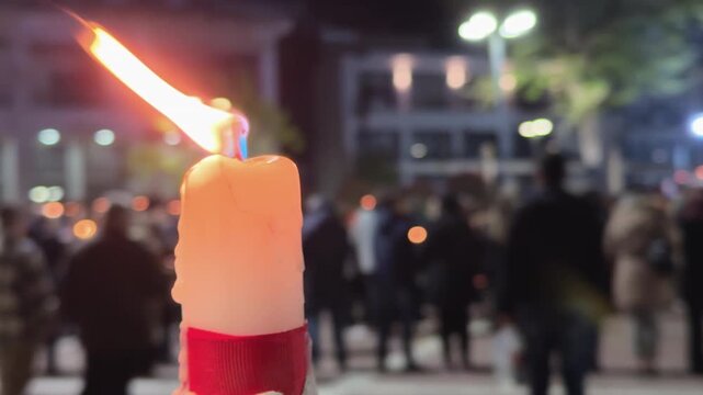Burning candle in sharp focus with blurred crowd holding lights in the background during an Easter nighttime outdoor gathering.