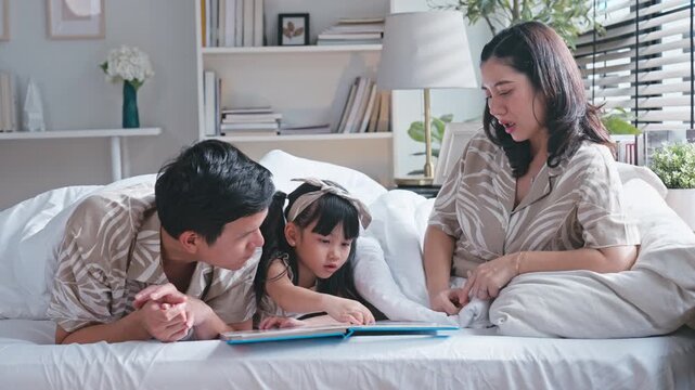 Asian family father and mother reading a storybook to their young daughter under a cozy blanket.