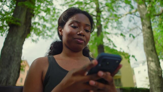 African American woman seated on park bench using smartphone and looking down at screen with calm and engaged expression creating natural and intimate lifestyle moment in outdoor environment
