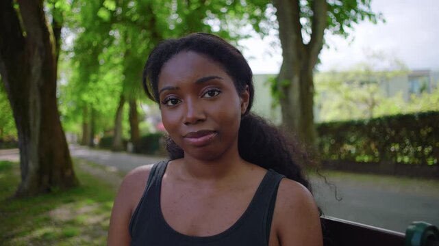 African American woman seated on park bench looking directly at camera with calm and neutral expression creating quiet and intimate outdoor portrait moment in natural environment