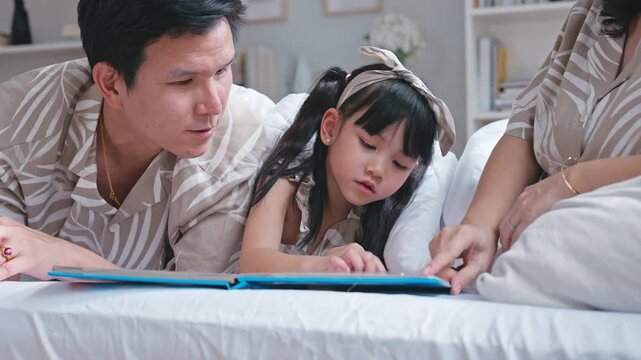 Asian family father and mother reading a storybook to their young daughter under a cozy blanket.