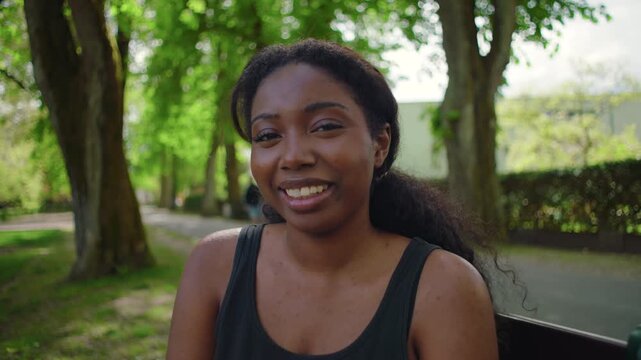 African American woman seated on park bench looking at camera and smiling warmly creating friendly and approachable outdoor portrait moment in natural environment