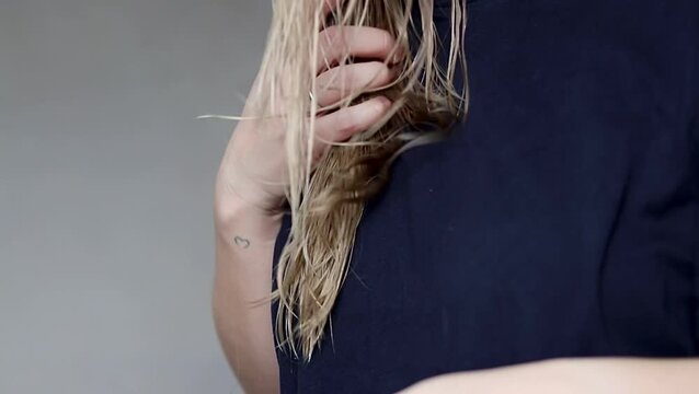 Female with long wet blonde hair gently combs fingers through strands while standing in a softly lit indoor space, showcasing hair care routine and personal grooming