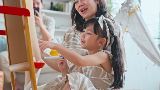 Asian family with daughter drawing on a canvas while mother helps and father captures the moment on a smartphone