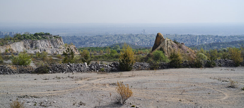 Wide view of an abandoned quarry with unique rock formations and sparse vegetation. Miskolc city stretches in the background under soft hazy light.
