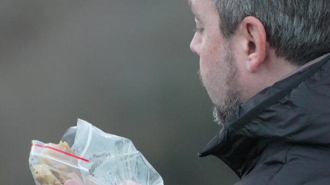 Man eating snack middle aged man with beard takes a bite of food from a plastic bag and looks at camera