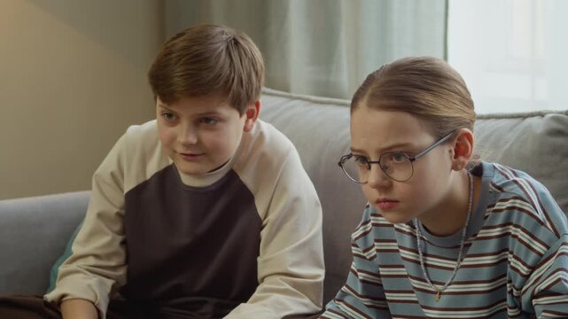 Handheld shot of focused gen Alpha siblings using controllers while playing video games sitting on couch in living room