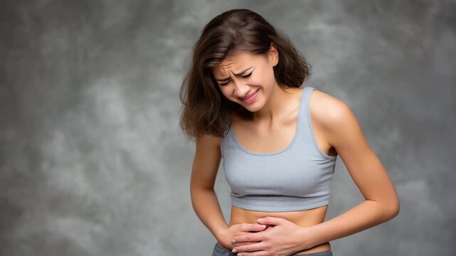 Indoor studio composition of a woman with abdominal pain, both hands covering stomach, body slightly turned to one side, expression of discomfort and illness, subtle shadows on gra