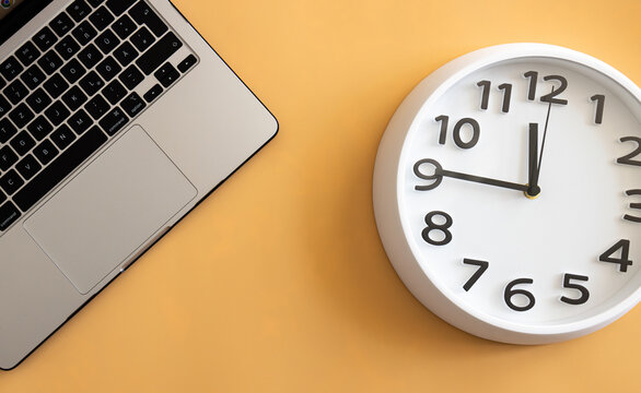 Modern office desk flat lay with laptop and wall clock on orange background for time management concept.