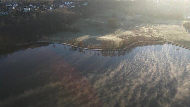 Aerial drone footage capturing early morning fog drifting over a lake in Stavanger, Norway during springtime. The scene features soft mist, calm water, and gentle morning light, creating a quiet and a