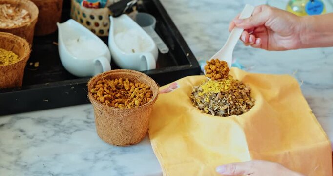 Hands filling yellow fabric sachet with dried aromatic herbs and flowers, marble countertop in hotel wellness workshop, spa lifestyle and aromatherapy diy activity