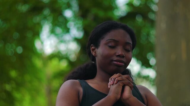 African American woman standing outdoors with hands clasped in prayer closing eyes then looking upward creating quiet and emotional moment of faith and reflection in natural environment
