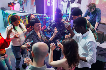Fototapeta premium Friends drinking alcohol while dancing and singing at nightclub party. Carefree young people enjoying beverages, holding glasses and saying toast while clubbing on dancefloor