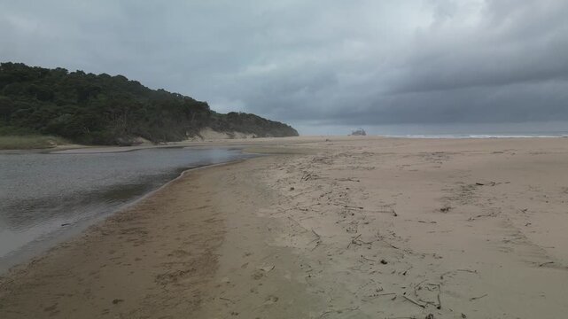 A low aerial view of a serene coastal landscape unfolds on a remote beach under a dramatic, cloudy sky, featuring a lagoon and dense coastal vegetation covering the hills.