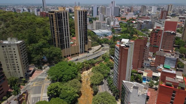 Drone tilt-up shot starting from the Cali River (Rio Cali) and lush riverbank trees, revealing the urban skyline, office buildings, and skyscrapers of downtown Cali under a bright, cloudy sky.