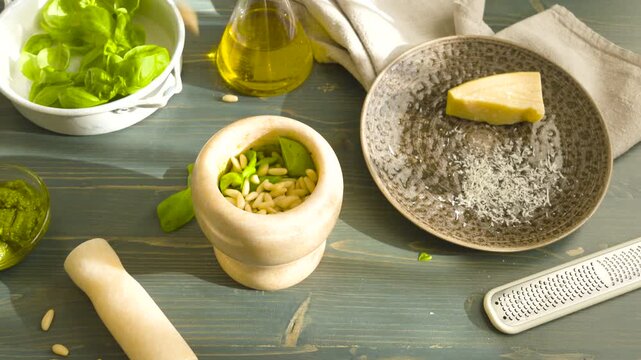 Adding pine nuts to marble mortar for traditional Genovese pesto. High angle shot featuring ingredients for authentic Italian sauce. Basil leaves, olive oil in glass decanter, parmesan cheese and