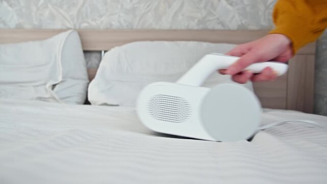Handheld vacuum cleaner removes dust from bed linens in bright bedroom setting