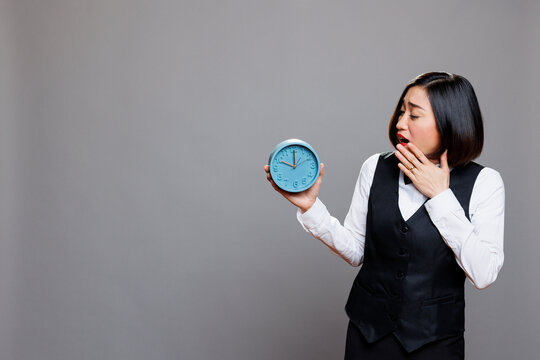 Asian waitress overslept and running late for work at cafe while yawning and looking at alarm clock. Restaurant sleepy woman employee wearing professional uniform showing time