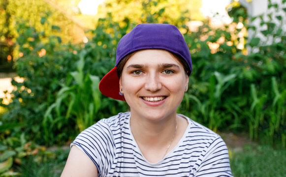 Close-up portrait of a smiling young woman wearing a purple baseball cap backwards and a striped t-shirt. Outdoor summer lifestyle concept with a blurred green garden background
