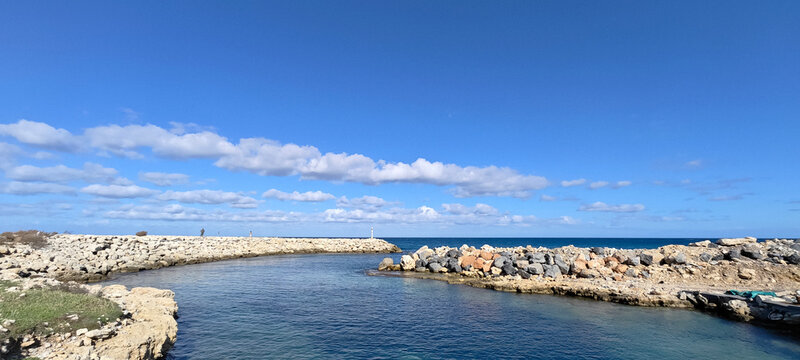 Panoramic view of a rocky breakwater protecting a calm coastal inlet. Deep blue Mediterranean waters under a bright sky with scattered white clouds, showcasing a peaceful harbor scene.
