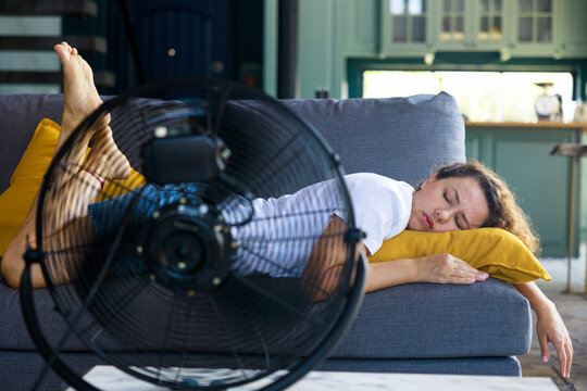 Woman sleeping on couch with fan during heatwave. Summer fatigue concept