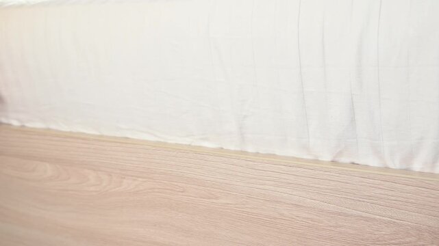 Hand tucks white bed sheet under wooden bed frame in bright bedroom setting