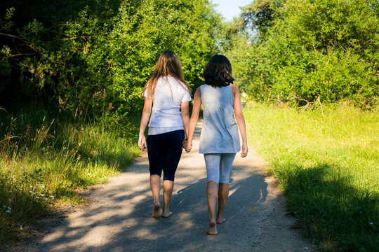 Two girls walking barefoot hand in hand along a rural path surrounded by greenery, summer countryside scene.
