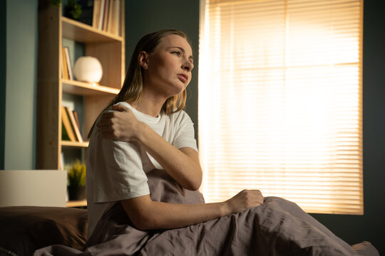 Woman sitting in bed, holding her shoulder in discomfort, feeling an injury or muscle ache after waking up, highlighting concepts of pain, exhaustion, and healthcare