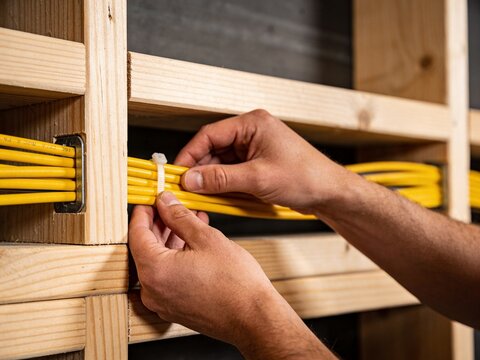 Hands of a person neatly bundling bright yellow electrical cables with a plastic zip tie inside a wooden wall framework, suggesting electrical installation work.