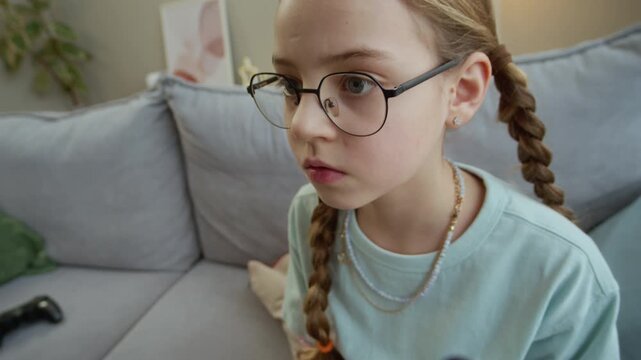 Tilt up shot of gen Alpha Caucasian girl using controller while playing video games sitting on sofa in living room