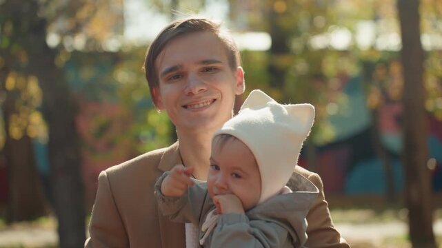 Young white father smiling with baby in park, baby chewing on teether, cozy beanie and warm coat, golden sunlight and mural backdrop, affectionate connection and family joy with marriage theme