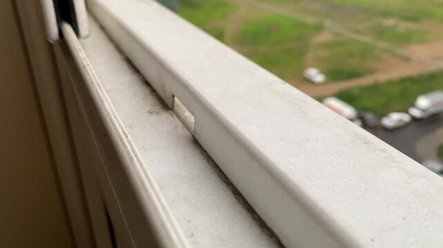 Closeup view of window slider opening process during day. Grass and cars seen outside, high floor at residential building. White window frame with dirt and dust