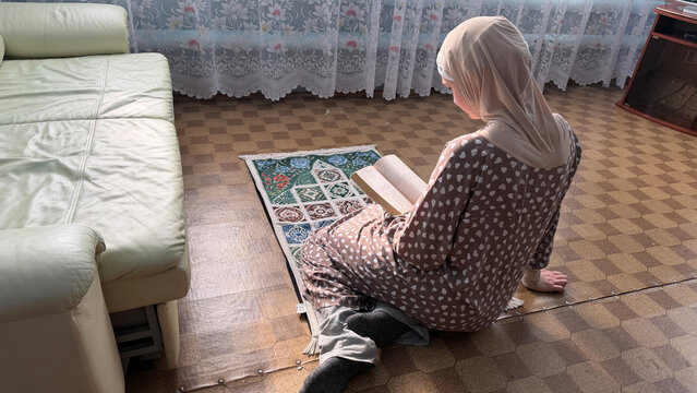 Muslim woman reading Quran on prayer rug at home