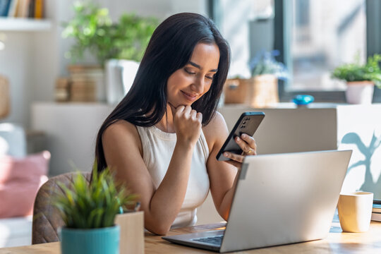 Beautiful woman using smartphone while working with laptop in the livingroom at home