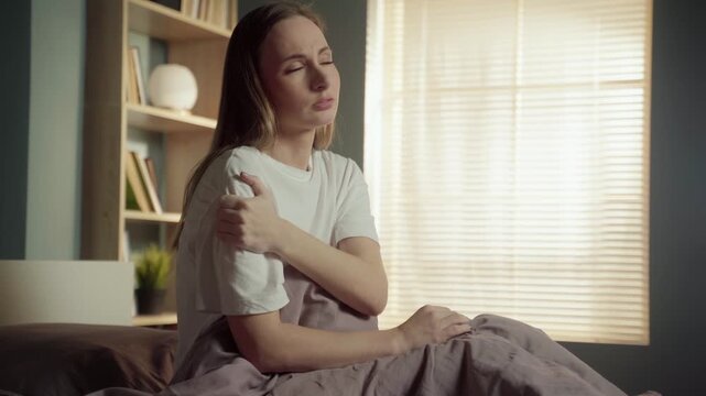 Young woman sitting in bed in the morning, feeling sharp pain and stiffness in her shoulder and neck after sleeping, massaging her sore joint with a pained expression on her face