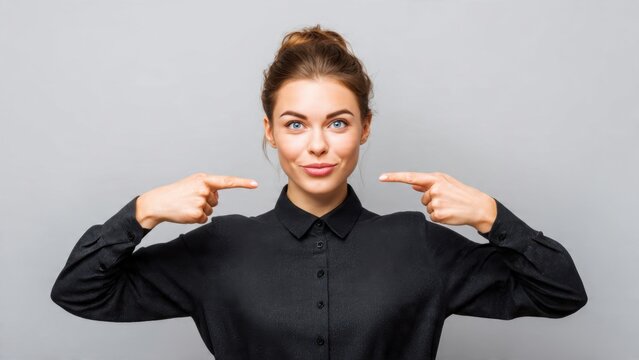 Confident young woman pointing to herself with both index fingers, making a choice or expressing self-affirmation against a grey background.