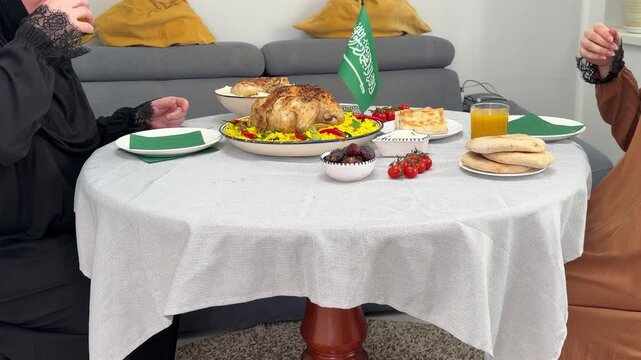 Two Muslim women in hijabs share traditional Middle Eastern meal. They sit at table with roasted chicken, rice, dates, and flatbread. Saudi Arabian flag decorates festive family dinner at home
